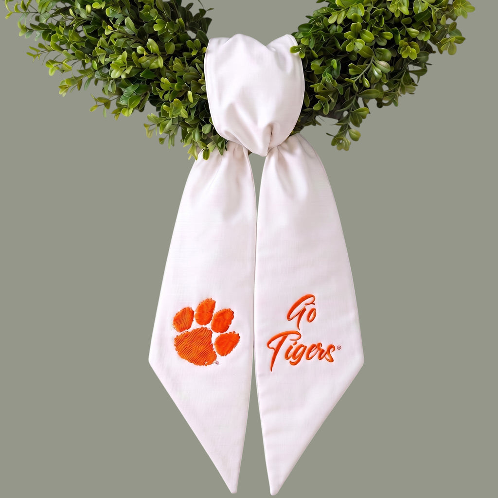 The Matthew John Collective white Clemson wreath sash with orange paw print and Go Tigers embroidery on green foliage