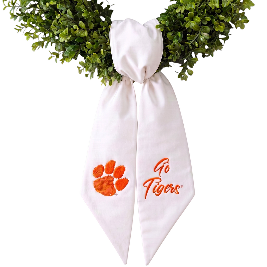 The Matthew John Collective white Clemson wreath sash with orange paw print and Go Tigers embroidery on green foliage