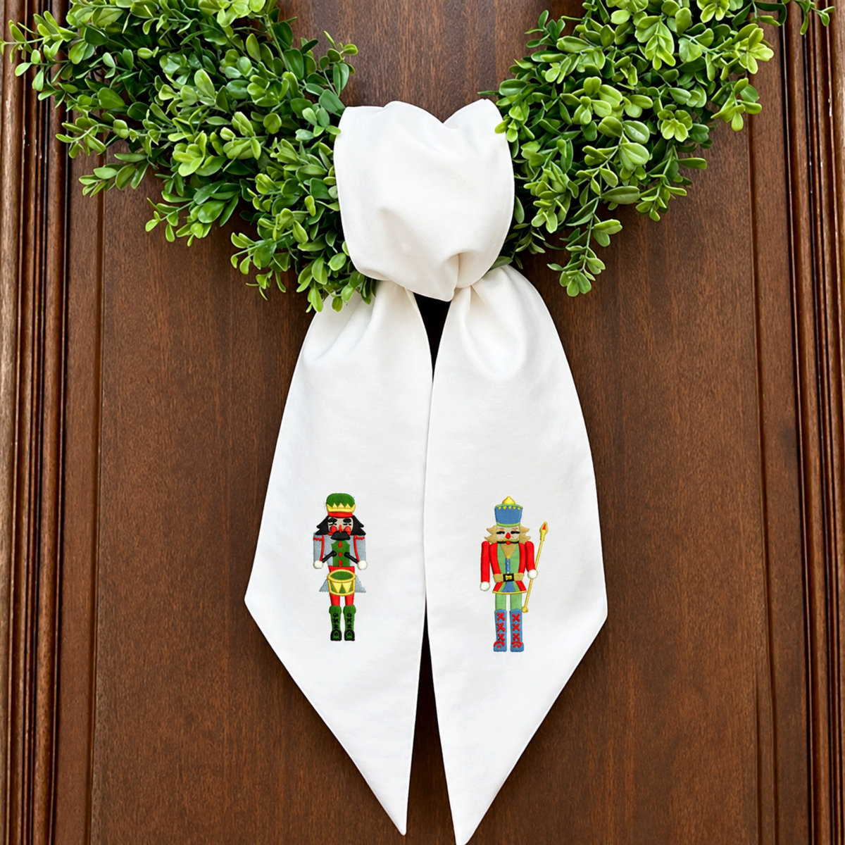 The Matthew John Collective white cotton canvas wreath sash with embroidered nutcracker soldiers on a green wreath.